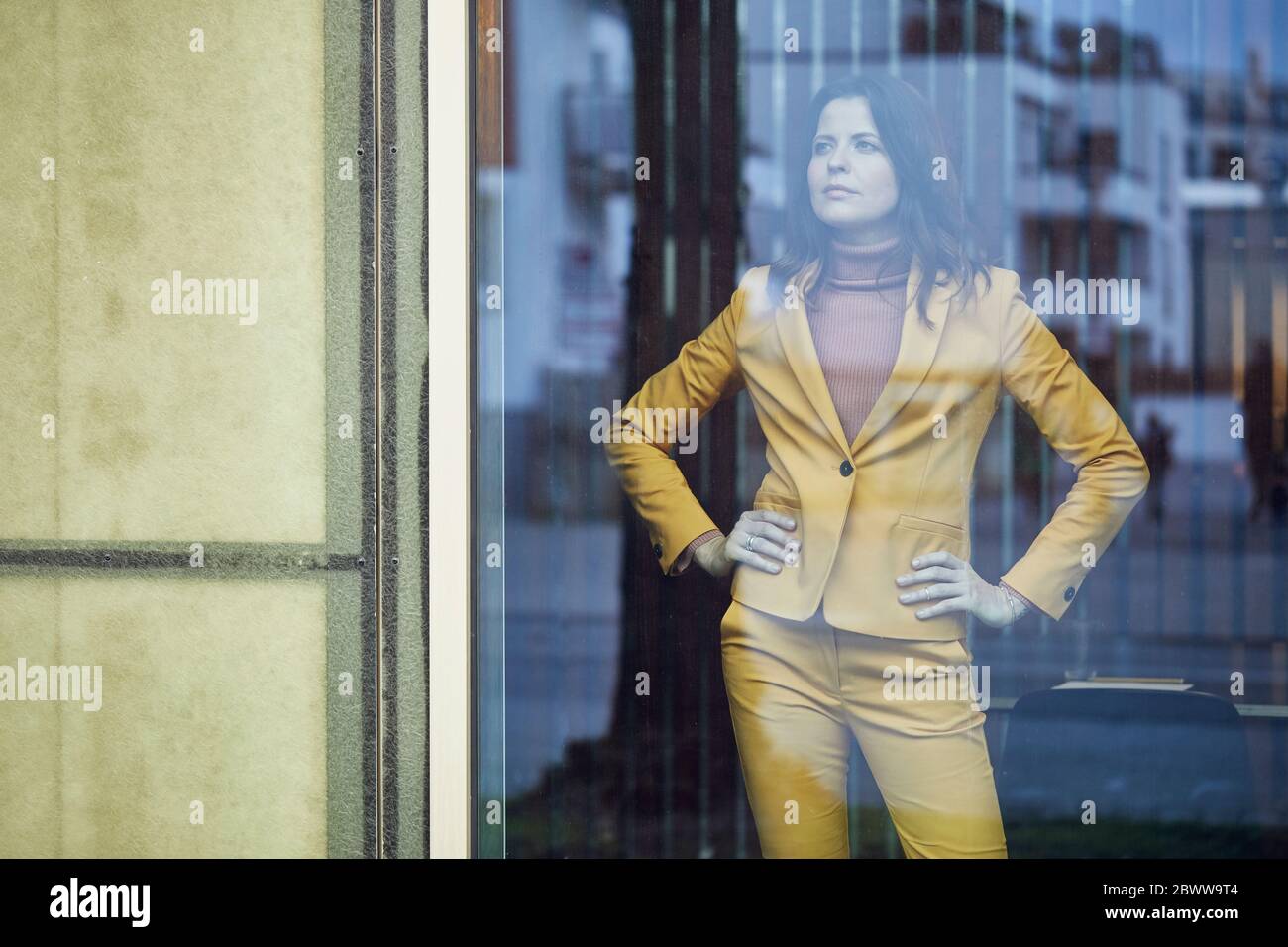 Confident businesswoman wearing yellow suit standing behind windowpane Stock Photo