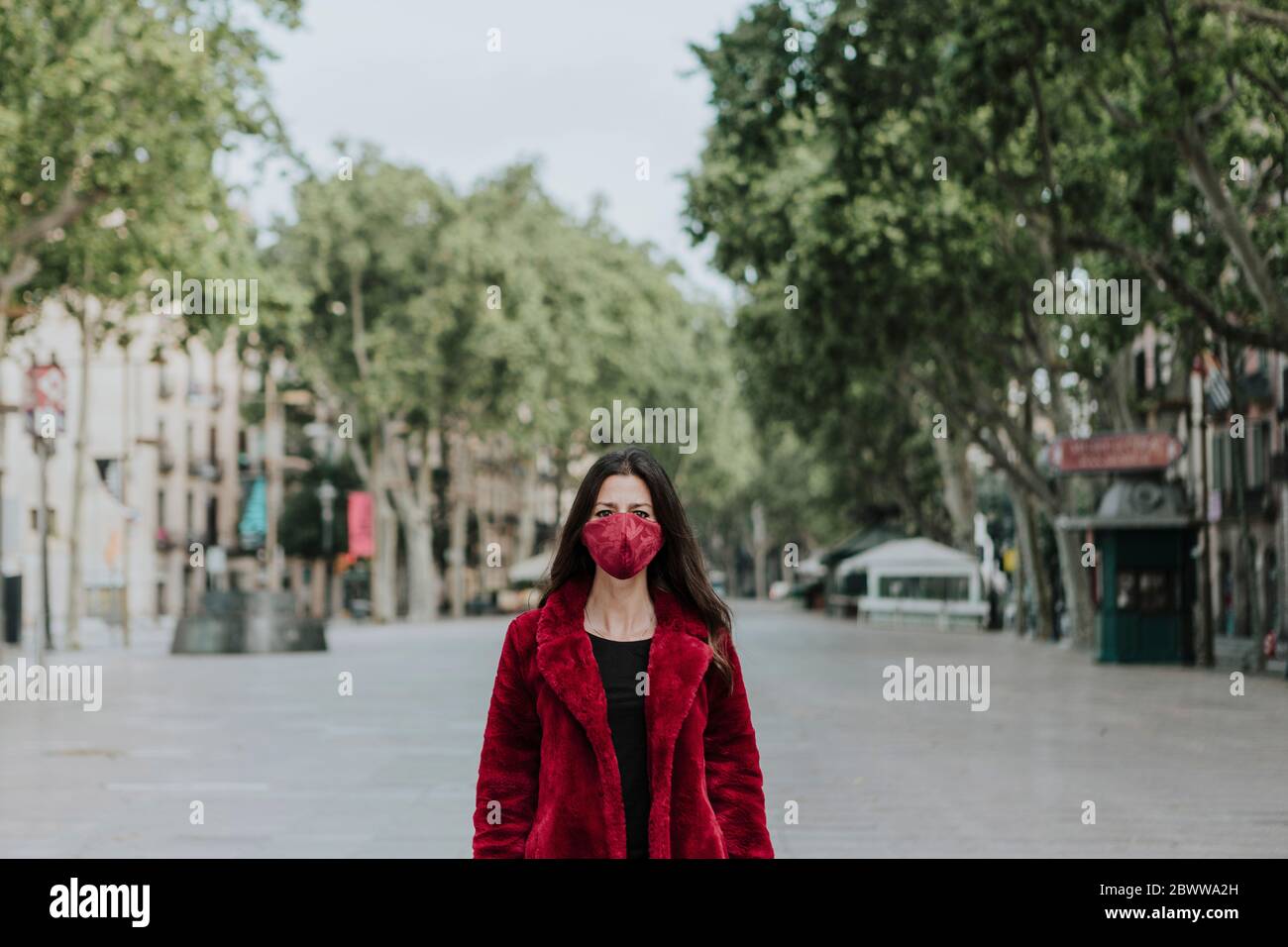 Portrait of woman wearing red face mask and jacket standing on empty street in city, Barcelona, Spain Stock Photo