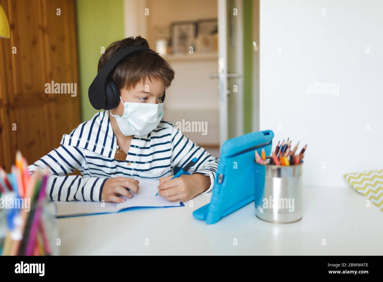 Boy doing homeschooling and writing on notebook, using tablet and headphones, wearing mask at home during corona crisis Stock Photo