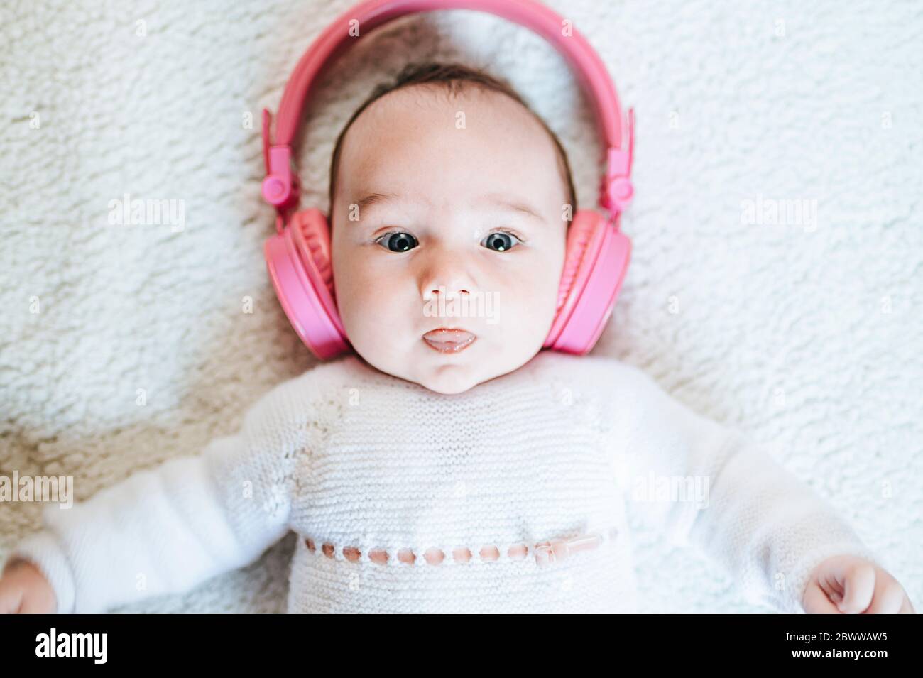 Portrait of baby girl with oversized pink headphones lying on blanket sticking out tongue Stock Photo
