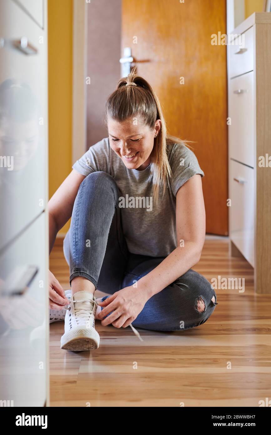 Smiling woman tying shoelace while sitting on hardwood floor at home Stock Photo