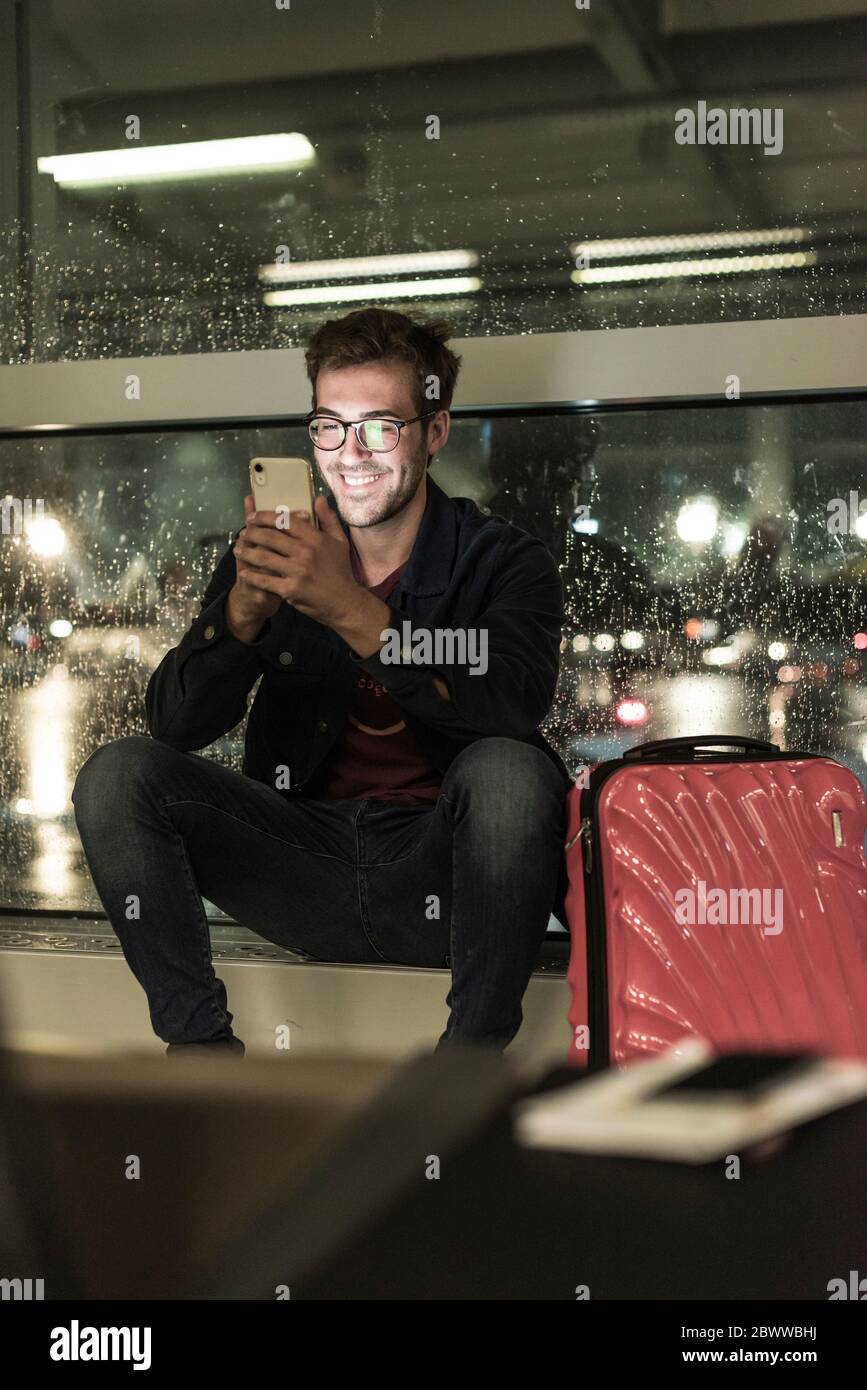 Smiling young man with suitcase sitting at rainy window using smartphone Stock Photo