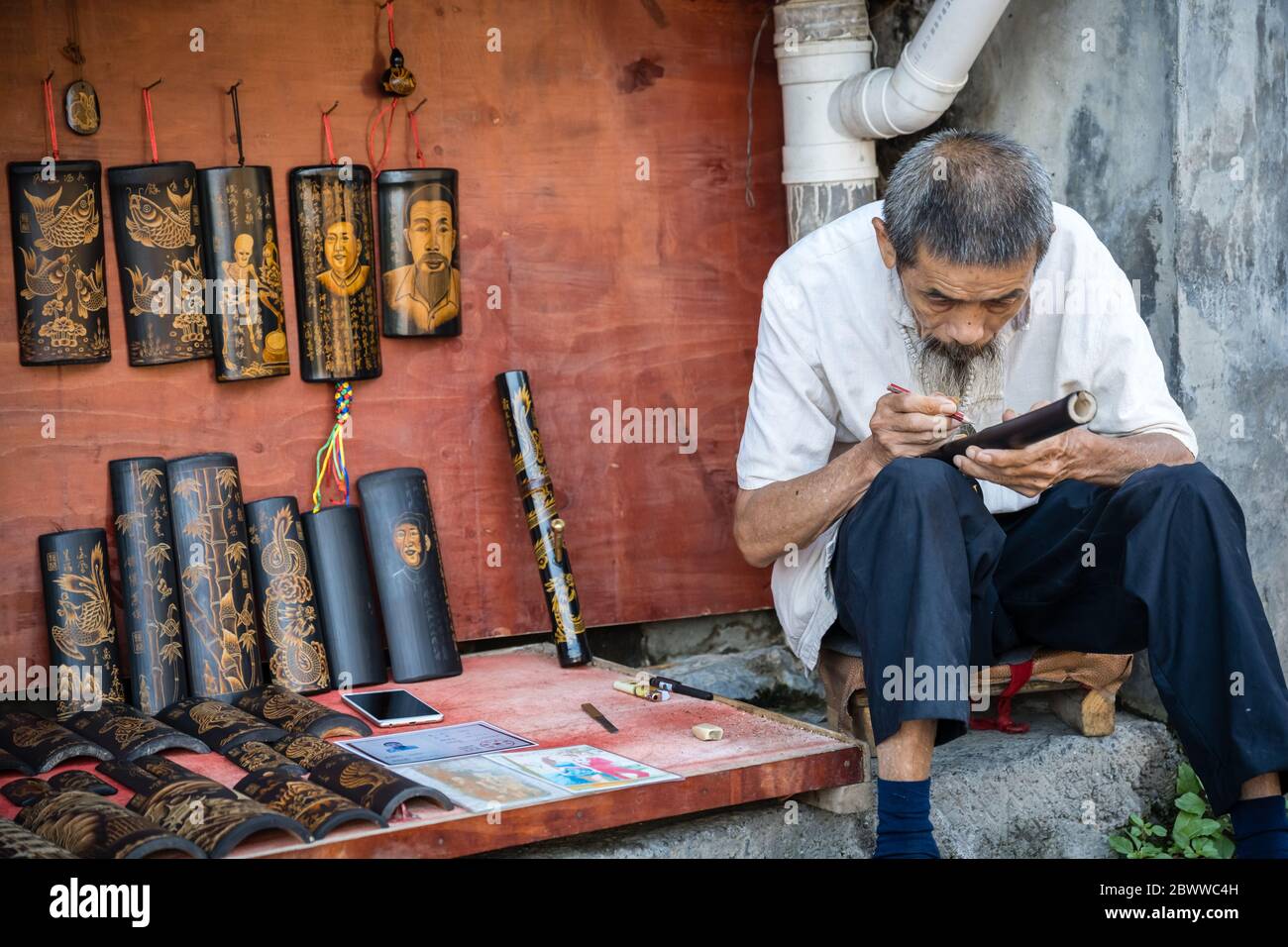 Feng Huang, China -  August 2019 : Old chinese artist man sitting on the street pavement and focusing on sketching picture drawn on a bamboo with penc Stock Photo