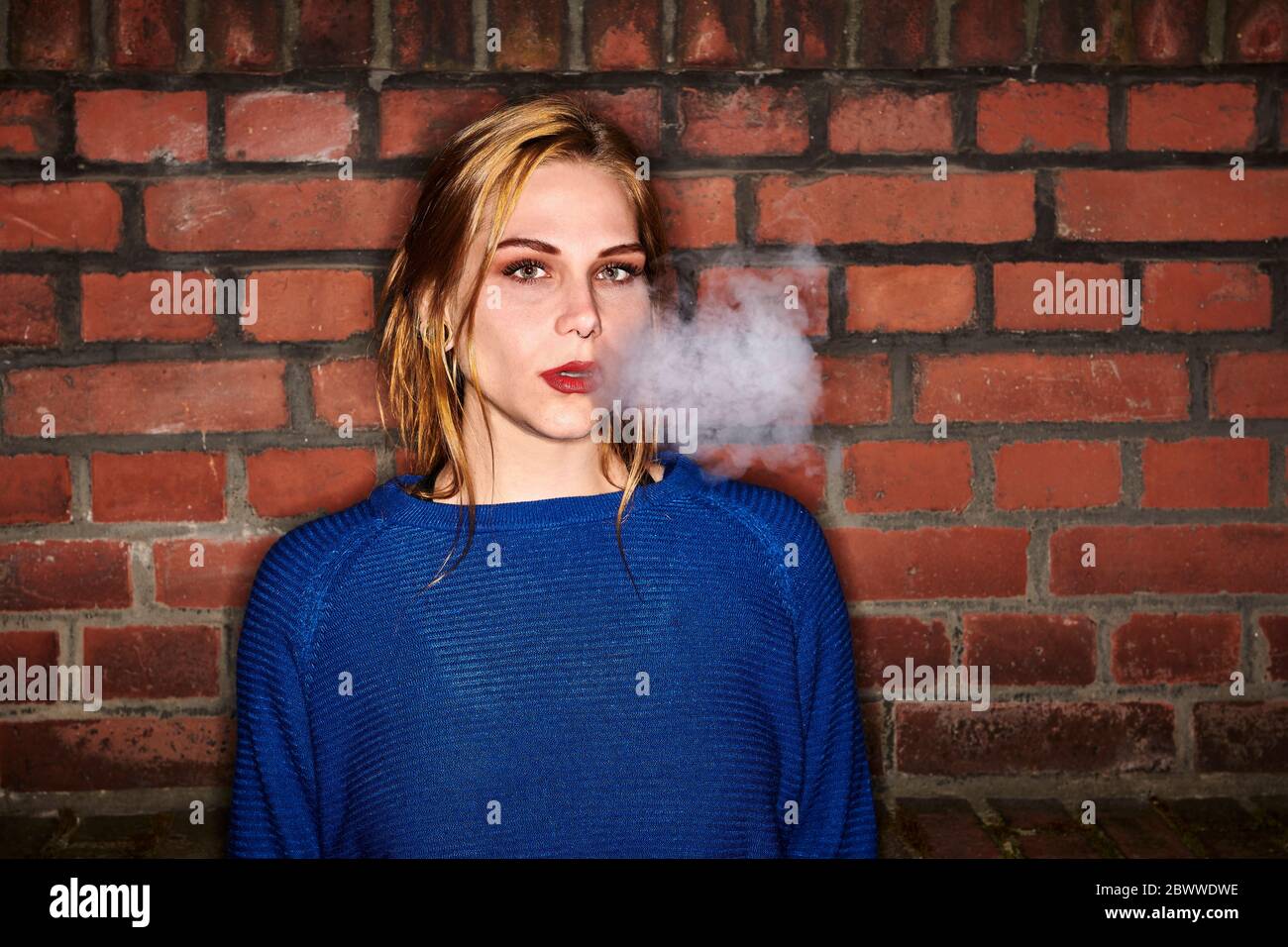 Portrait of young woman exhaling smoke against brick wall at night Stock Photo