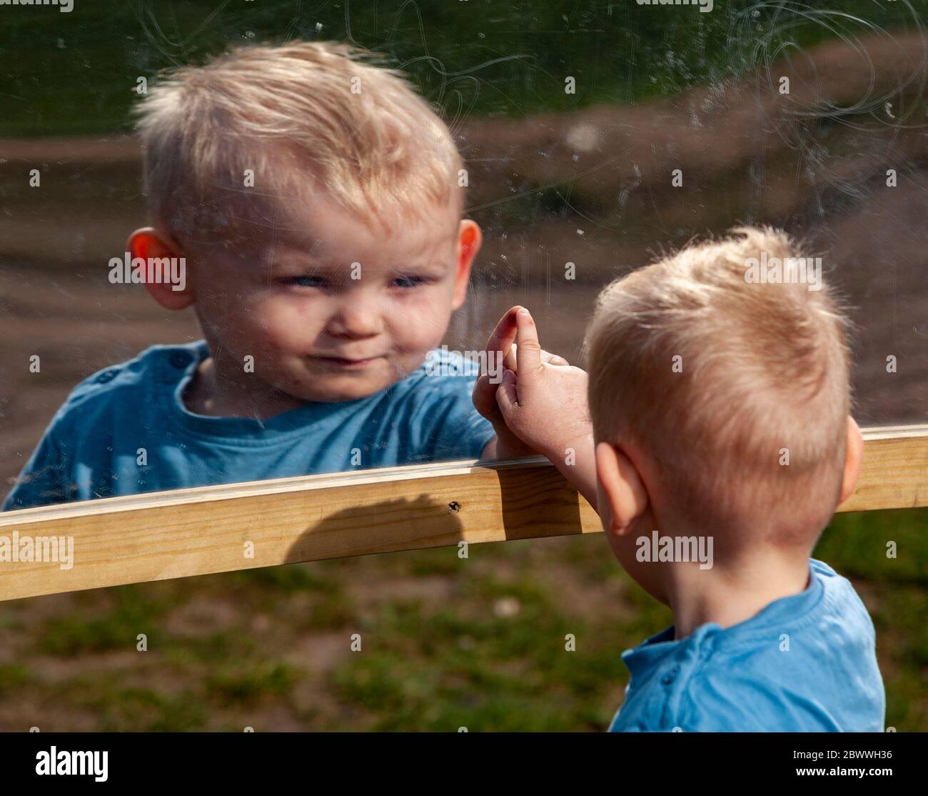 2 year old toddler boy pointing at his reflection in a playground mirror UK Stock Photo