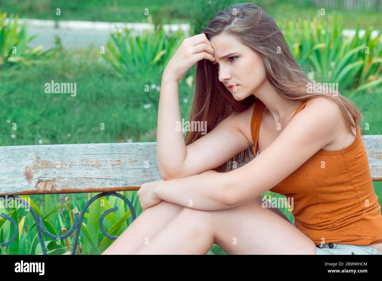 Thoughtful and sad girl. Portrait of one woman looking down sadly thinking sitting on a bench in a green park outdoors background in summer. Horizonta Stock Photo
