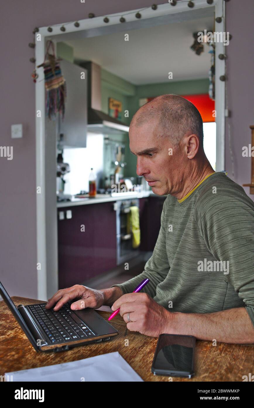 Man working at home at kitchen dining room table with laptop computer and mobile phone. UK Stock Photo