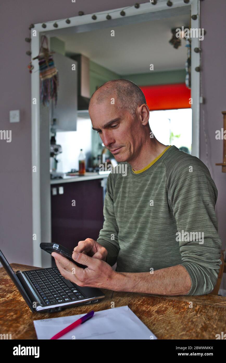 Man working at home at kitchen dining room table with laptop computer and mobile phone. UK Stock Photo