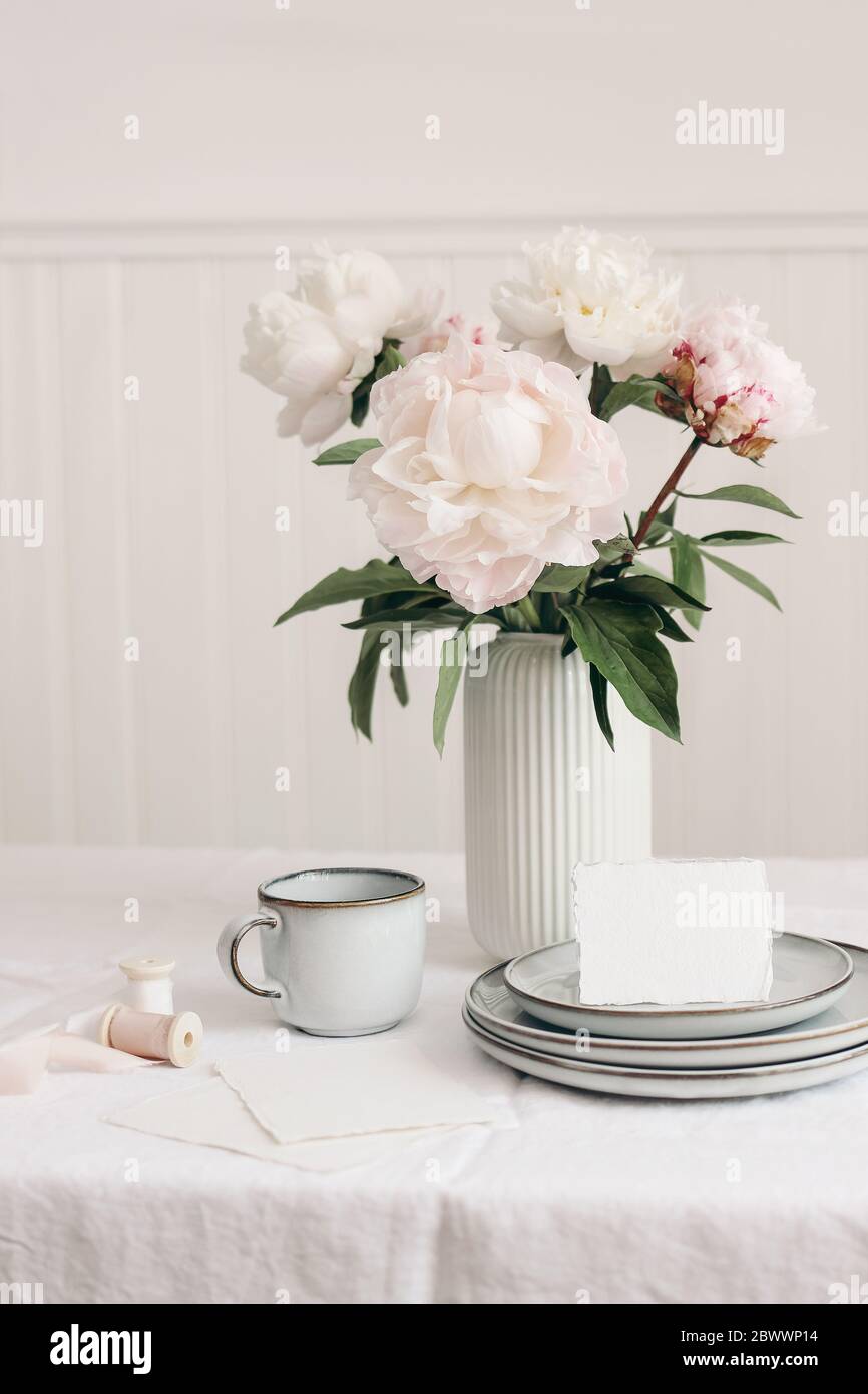Wedding still life scene. Business, place card mockup scene, marble tray, cup of coffee an pink peony flowers in vase. White linen table cloth. Floral Stock Photo