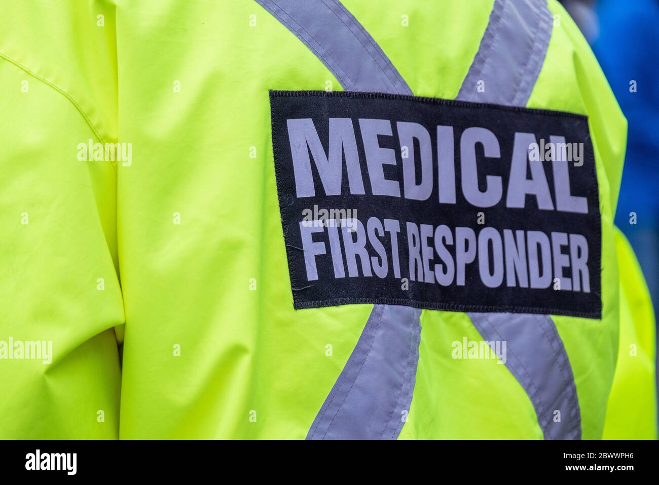 A bright yellow medical first responder uniform being worn by a large male in a crowded street.The coat has a grey reflective cross across the back. Stock Photo