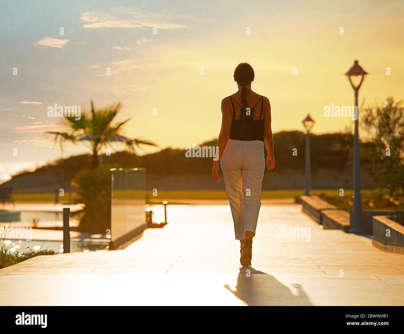 Back view of female silhouette going on evening sunset background of resort hotel. Woman walking by tropic street way with palms and mountain Stock Photo