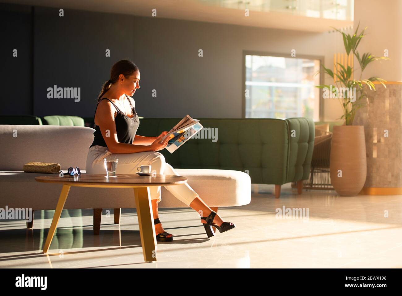 Young adult woman reading magazine on sofa in light modern interior of hotel reception, office building hall, waiting area background Stock Photo