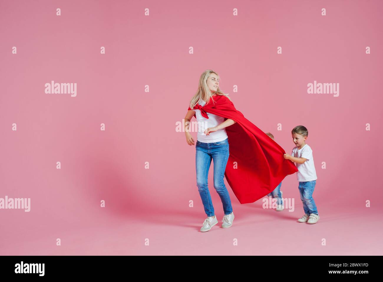 Mom is a superhero. Fun family, a young blond woman in a red Cape and her son, Sons follow mom pink background. Stock Photo