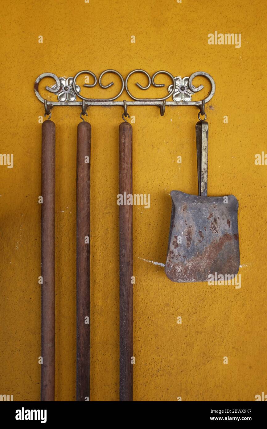 Metallic hand shovel hanging on a yellow wall next to three wooden sweeper handles Stock Photo