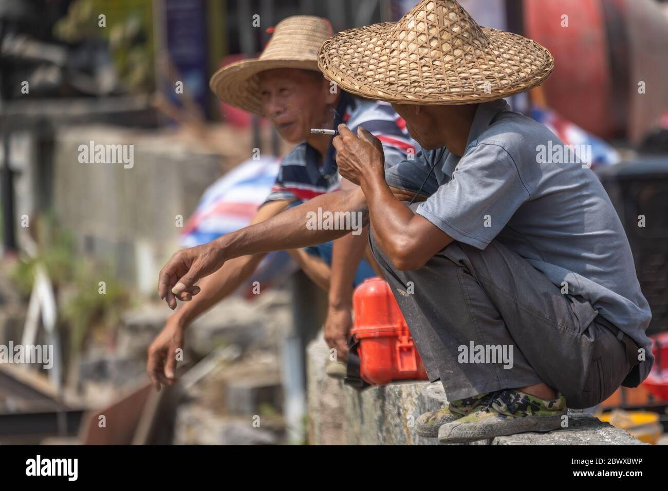 Yangshuo, China - August 2019 : Two Chinese men wearing traditional wicker conical hats smoking cigarettes and chatting during work break Stock Photo