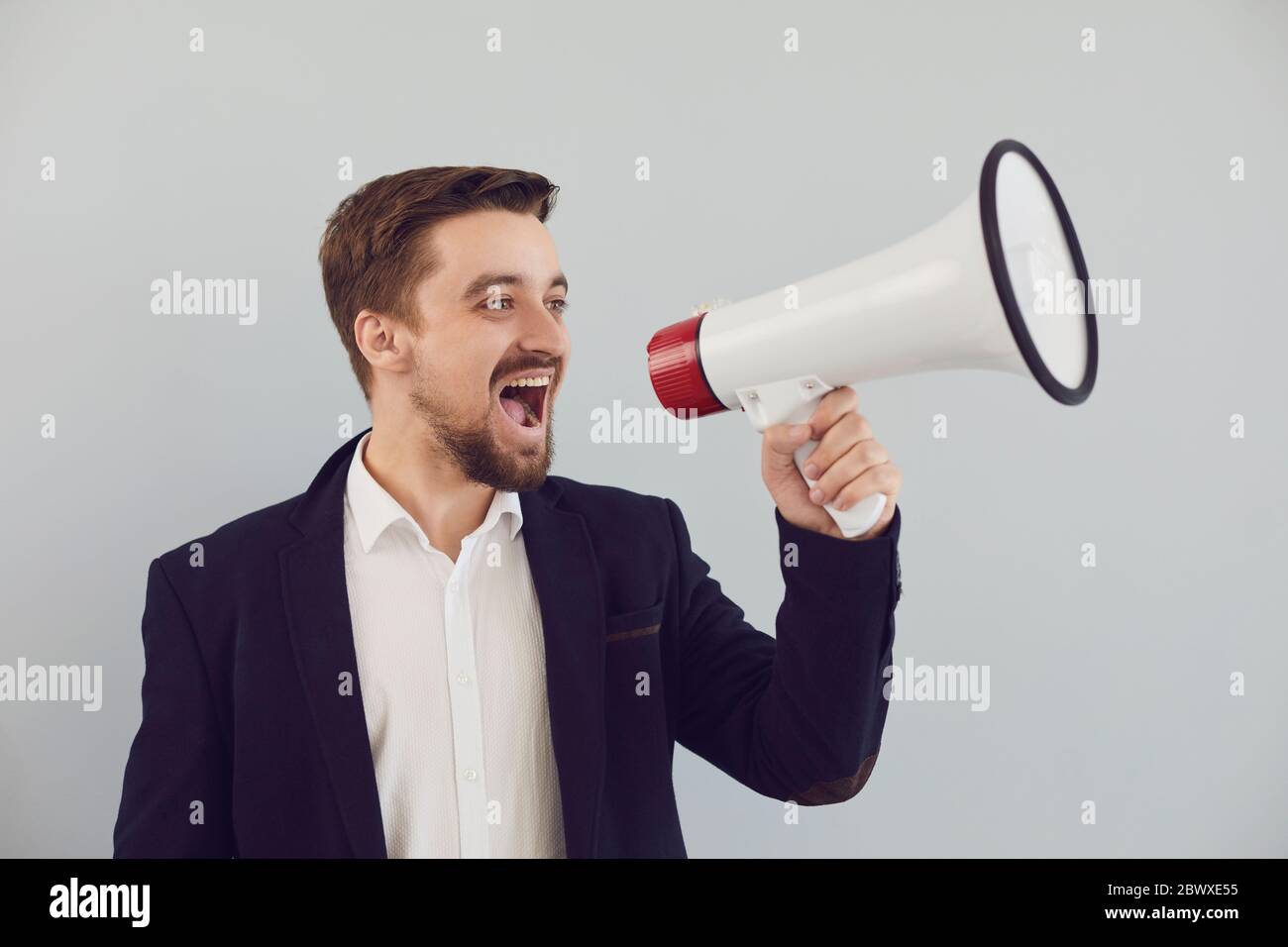 A man in a jacket shouts in a bullhorn on a gray background. Stock Photo