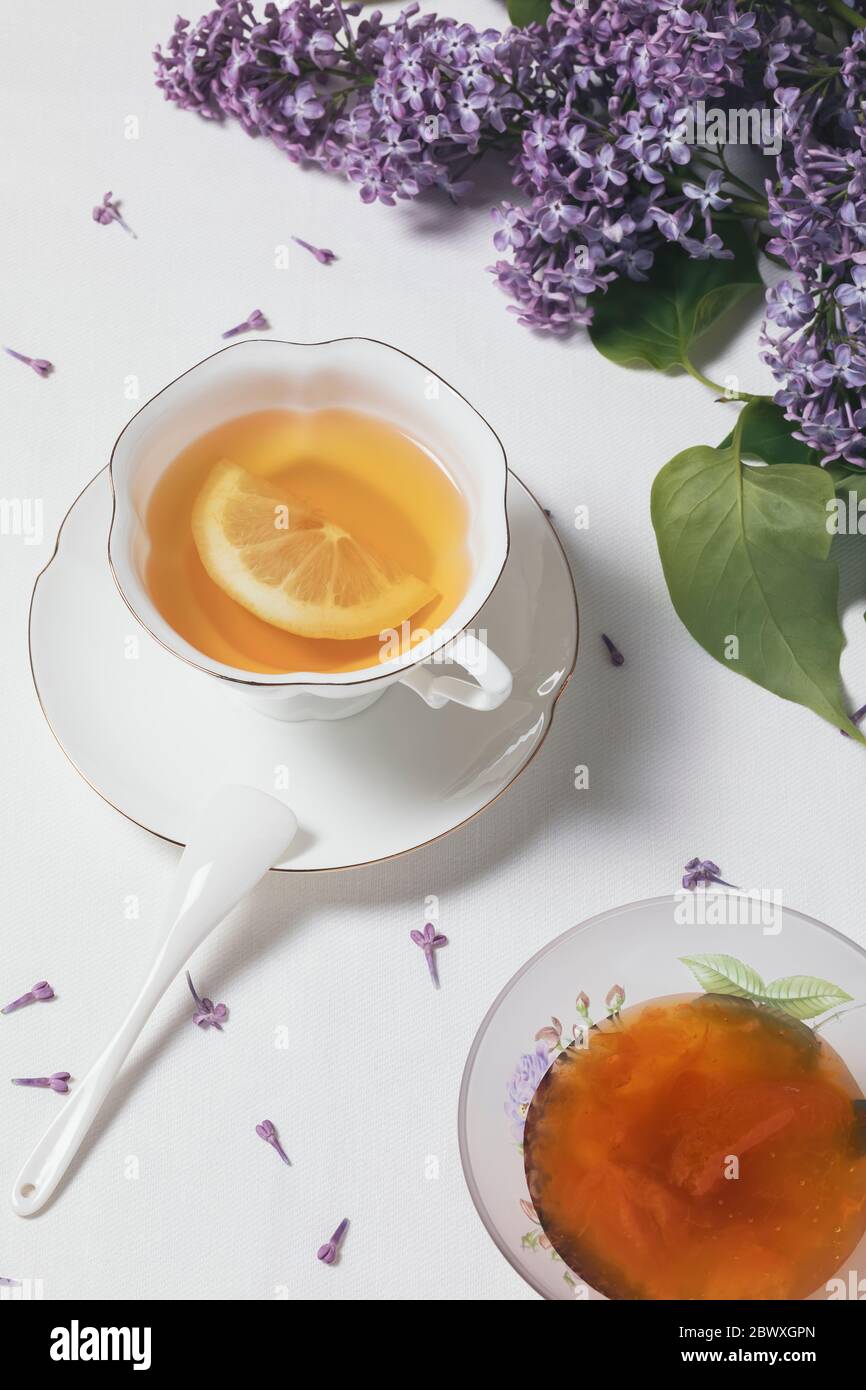 Bone china cup of tea with lemon, bowl with apricot jam and a vase with spring flowers (lilac) on white background Stock Photo
