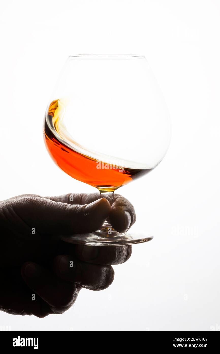 Man's hand holding snifter with cognac. Splash of a drink in a glass. White bright background. Stock Photo