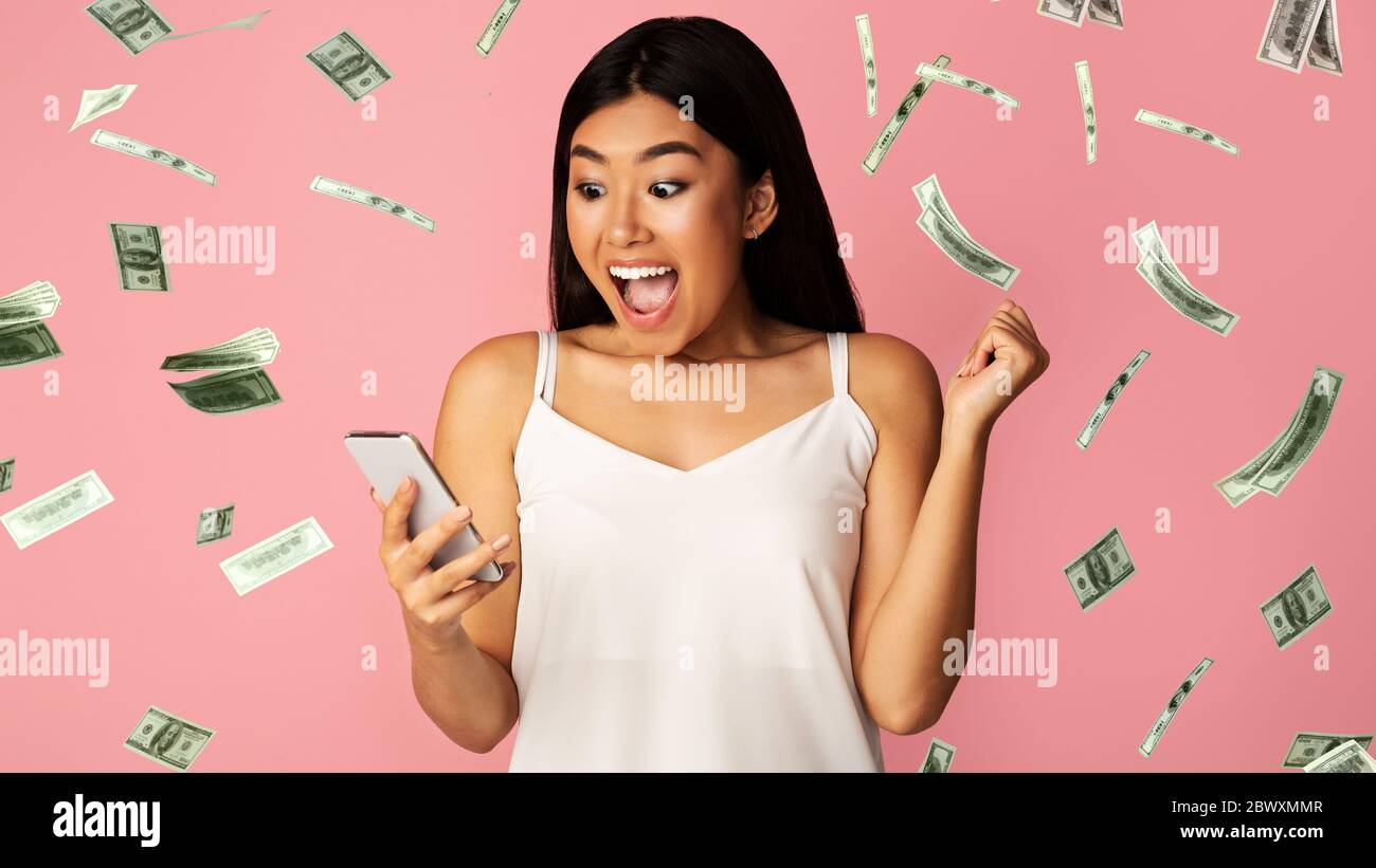 Big Win. Asian Girl Celebrating Victory With Smartphone Under Dollar Money Shower Stock Photo