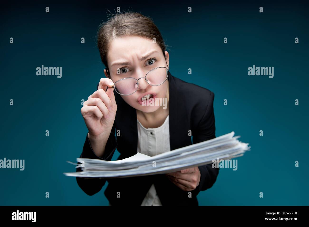 A young woman puts glasses on her nose and looks in camera with angry surprise. Director dissatisfied with paperwork or errors in them. Teacher scream Stock Photo