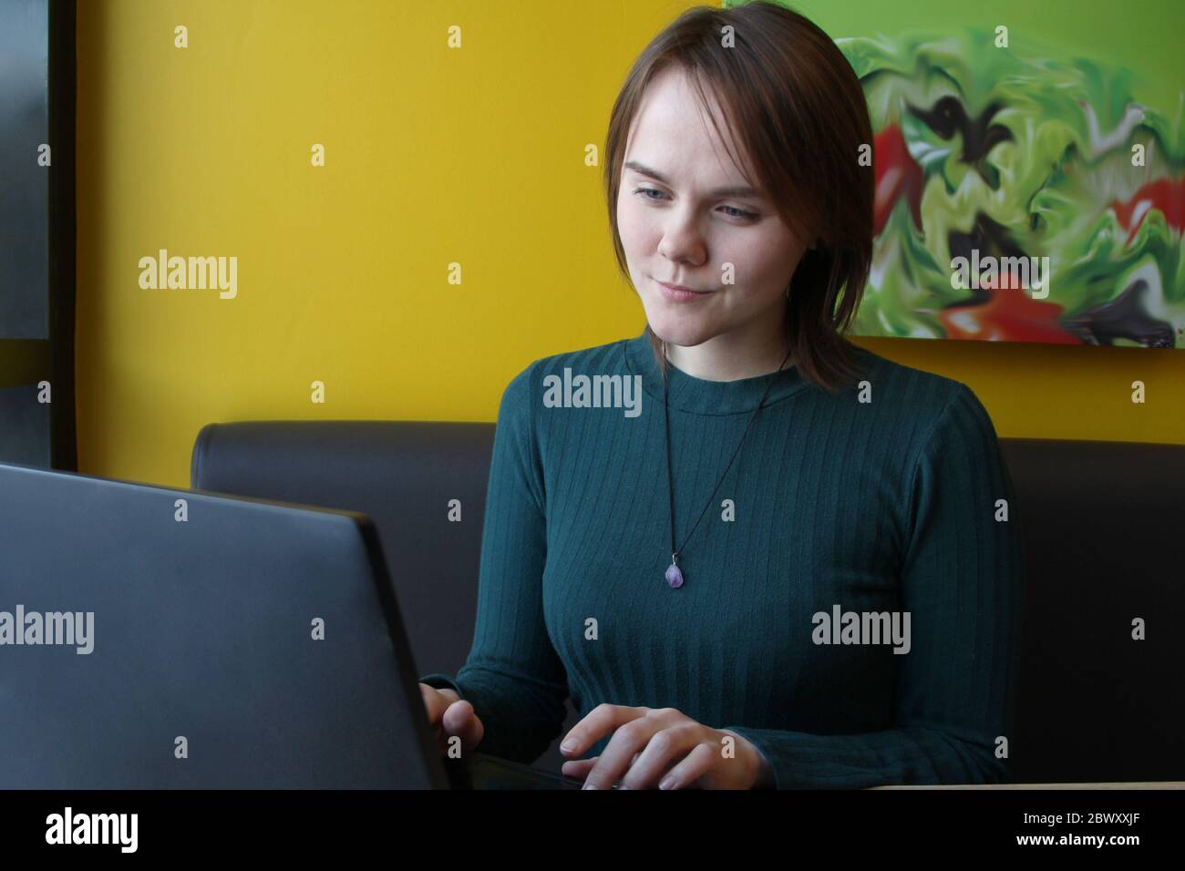 A girl with a pensive tense interested expression on her face sits, working at a laptop at a table in a cafe on a brown sofa against a yellow wall. He looks at the computer screen with narrowed eyes. Stock Photo