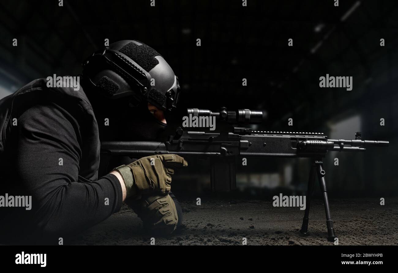 Photo of a soldier in helmet and ammunition laying and aiming sniper rifle profile view on dark background. Stock Photo