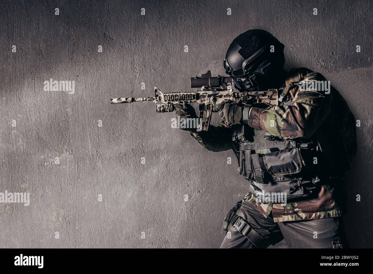 Photo of a fully equipped soldier in camouflage uniform standing and aiming rifle on grungy concrete wall background. Stock Photo