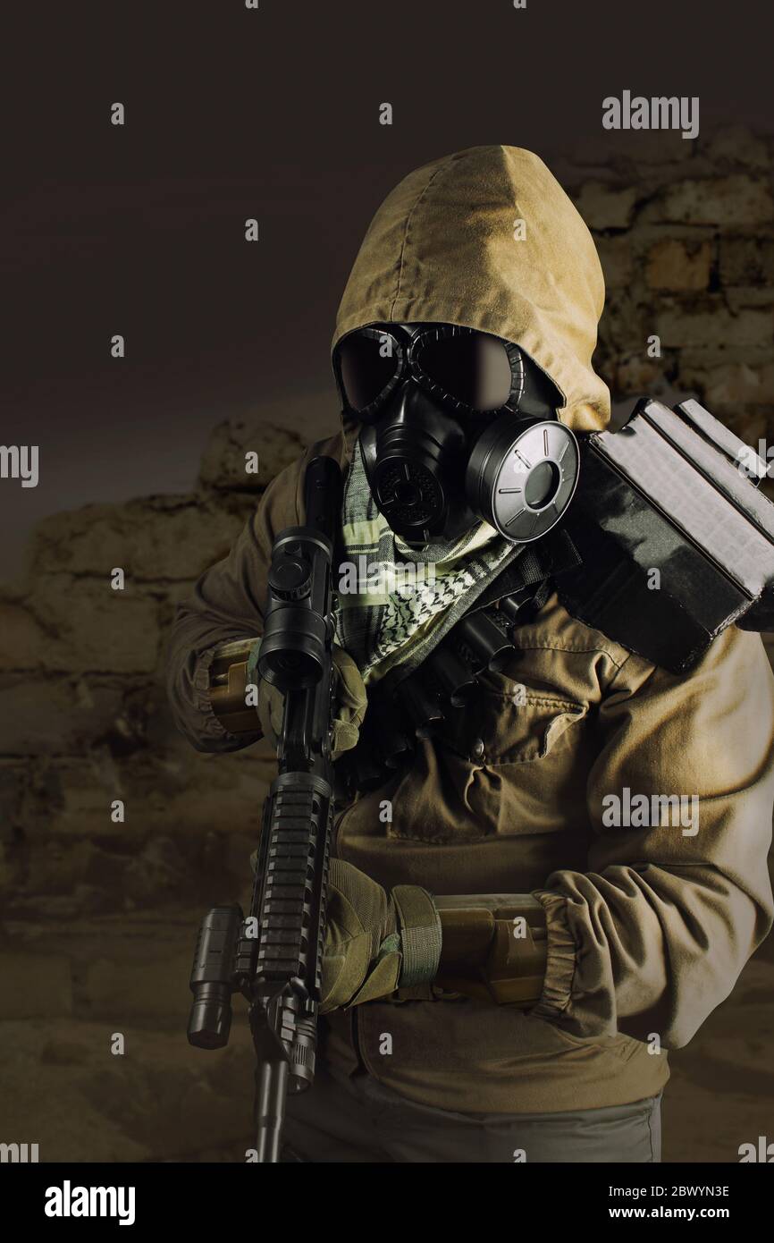 Photo of a desert post-apocalyptic soldier in tactical jacket, gas mask, gloves, rifle and armor standing on shaded wasteland background front view. Stock Photo