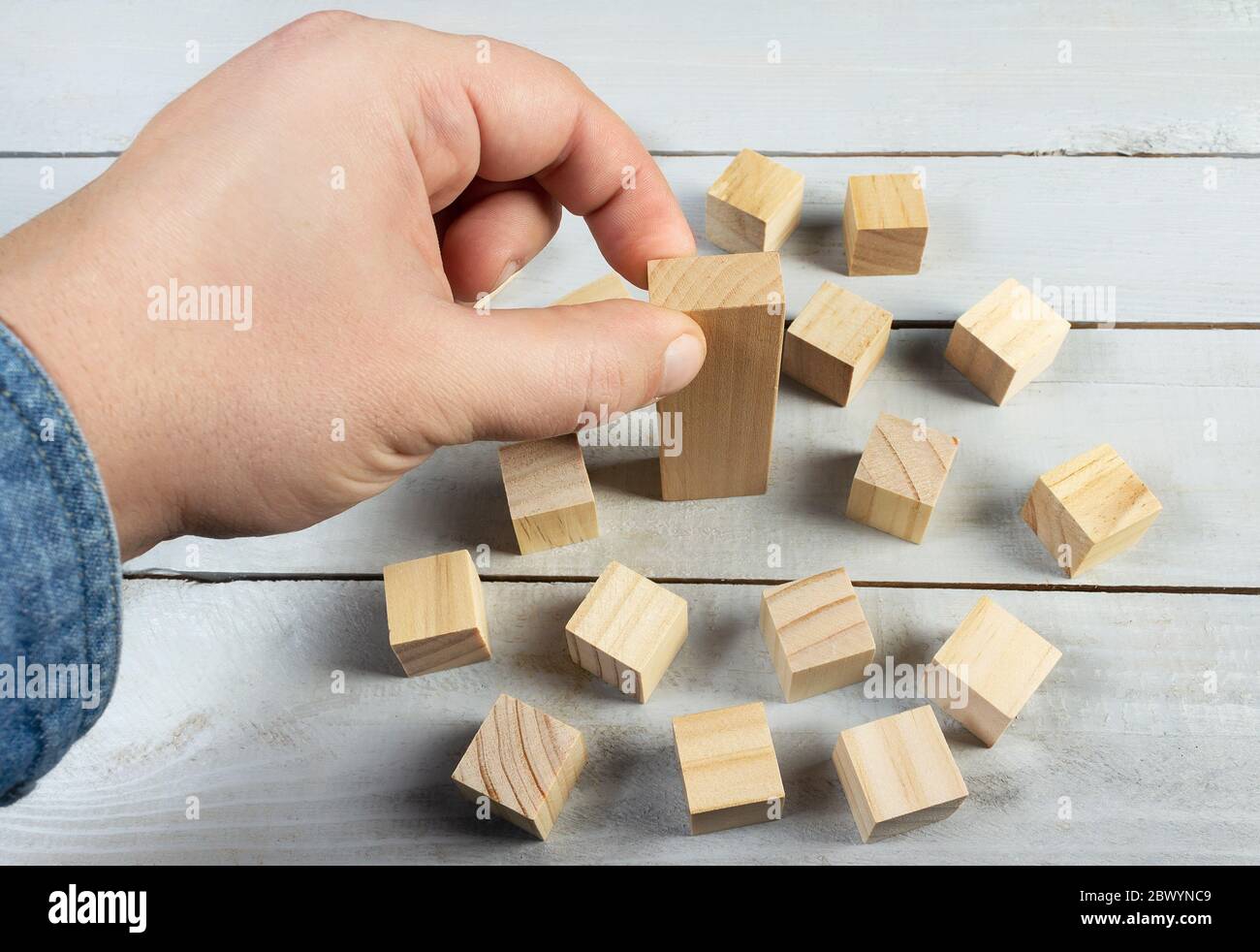 Concept photo of hand in jeans shirt putting a bigger wooden block in surrounding of a smaller ones on a table surface background. Stock Photo
