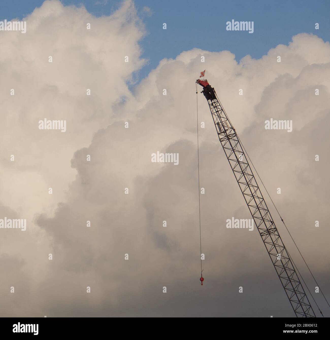 Tall construction crane boom stands with suspended hook and puffy cumulus clouds behind it. Metaphor for success, achievement, ambition, height. Stock Photo