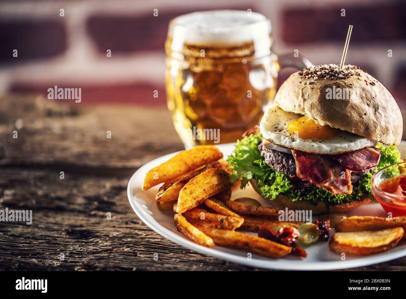 Burger with a sesame bun, fried egg and bacon, salad and potato wedges with a draft beer in the background Stock Photo