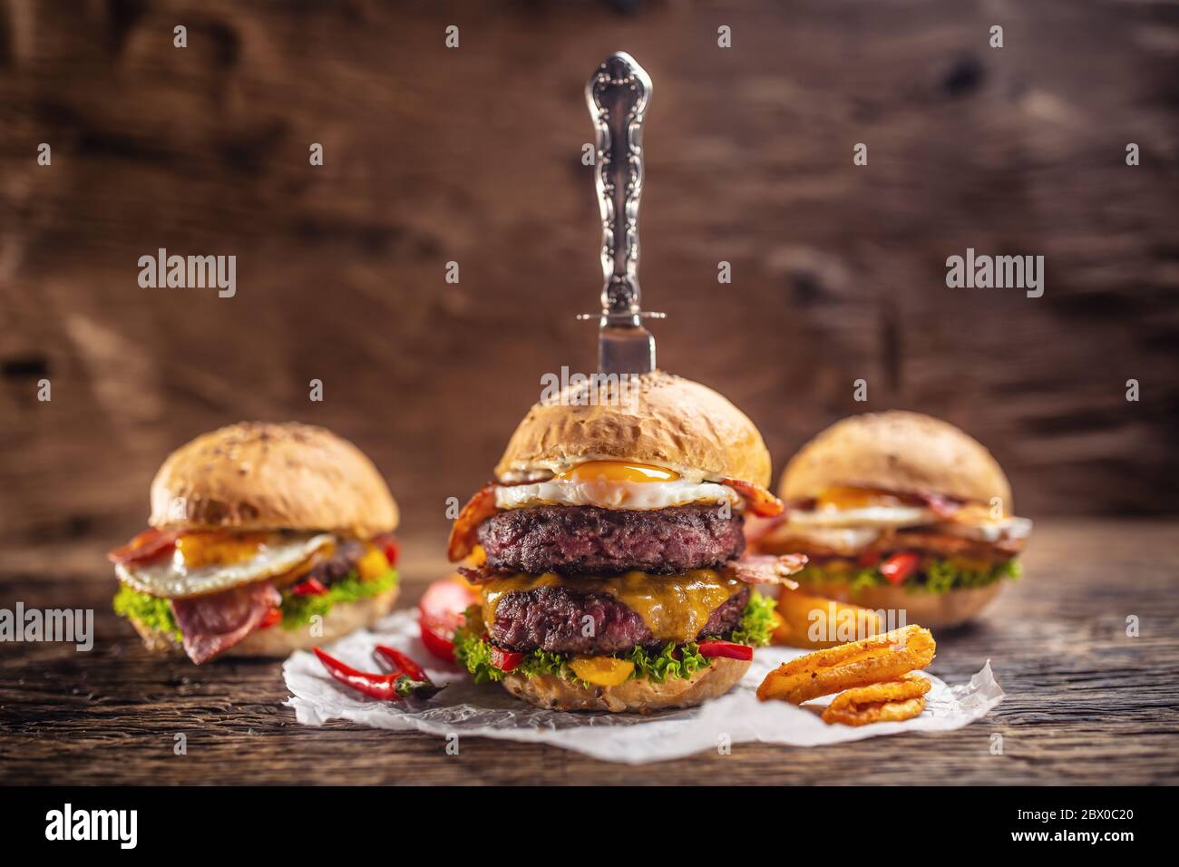 A knife-stabbed double beef burger with chips and chilli, more burgers in the back in a dark environment Stock Photo