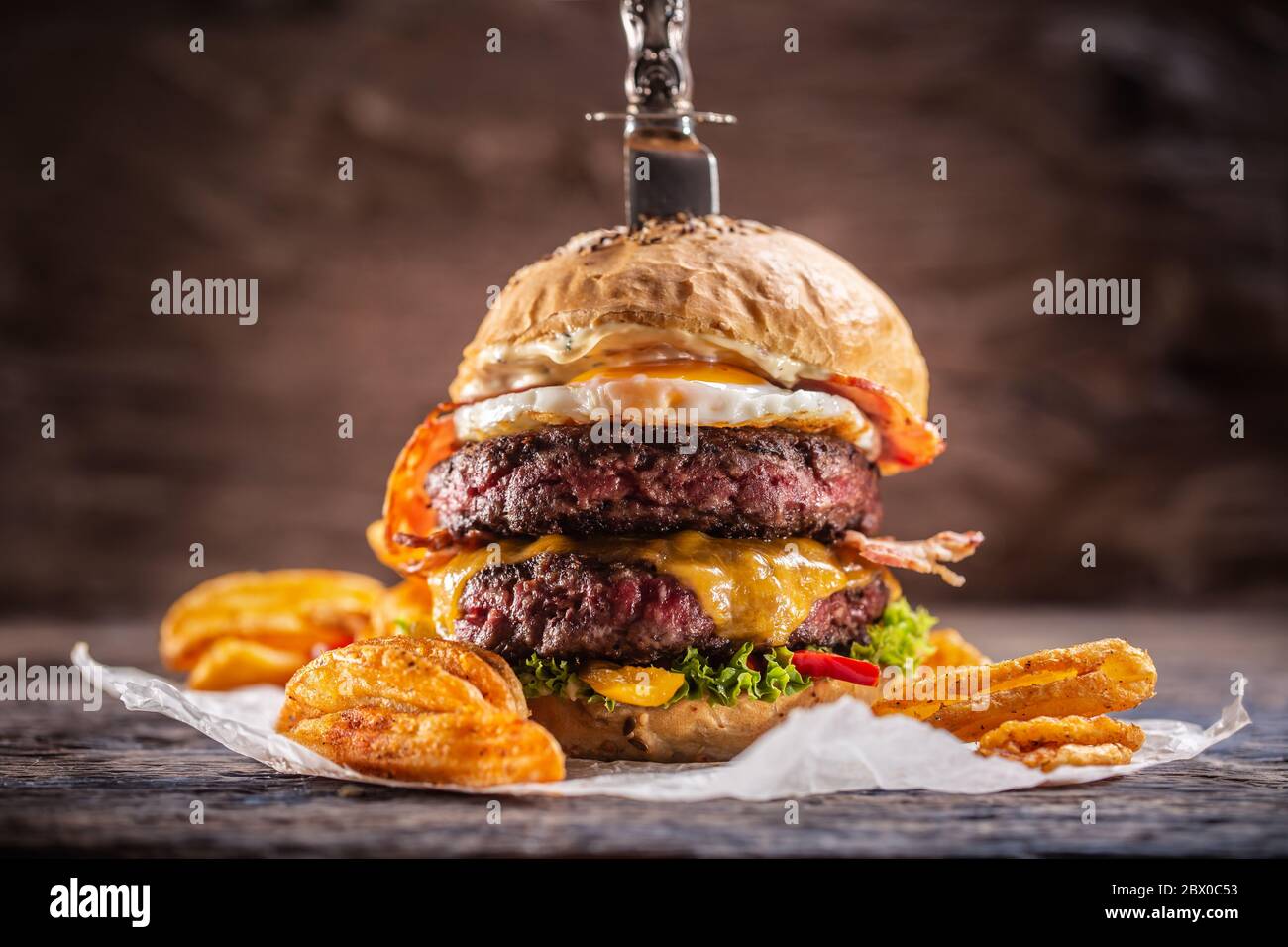 A knife-stabbed double-beef burger with melted cheese, fried egg and bacon, salad and paprika potato wedges Stock Photo