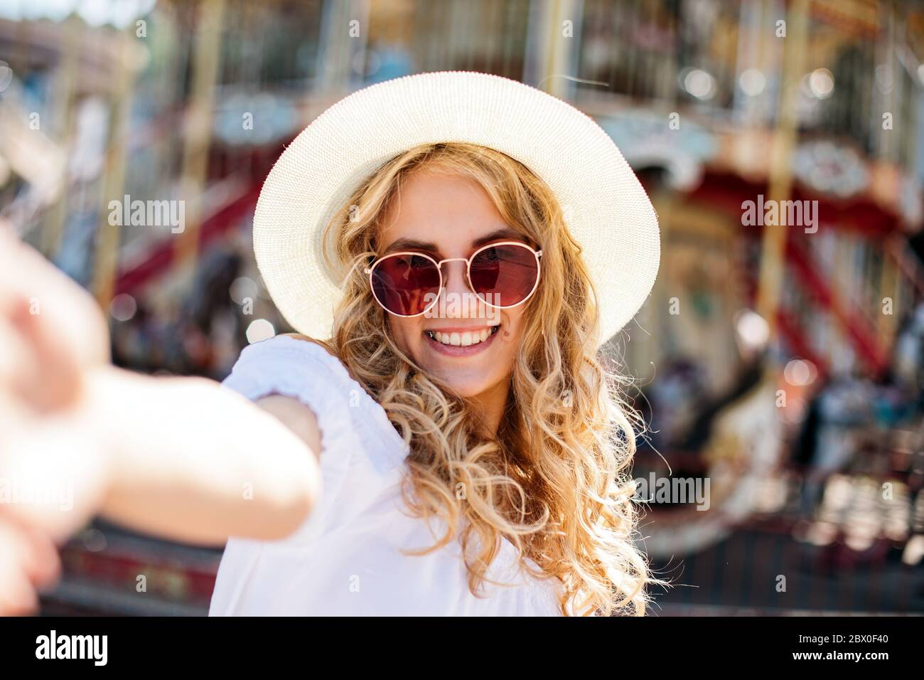 Joyful smiling girl in pink sunglasses making selfie in front of carousel. Outdoor portrait of cute stylish woman having fun in amusement park and tak Stock Photo