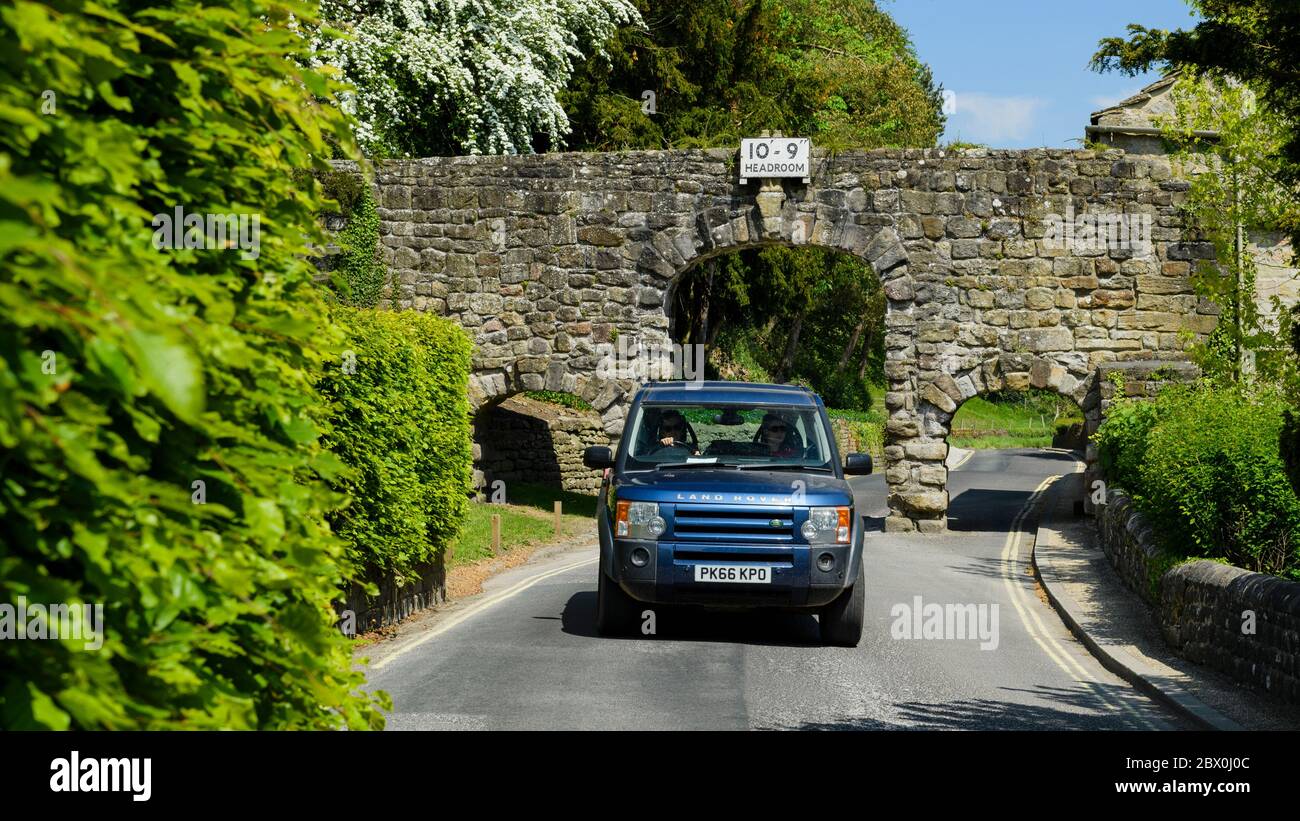 Car on scenic lane driving & low narrow stone archway (3 arches, headroom warning sign 10' 9'') - B6160, Bolton Abbey village, Yorkshire, England, UK. Stock Photo