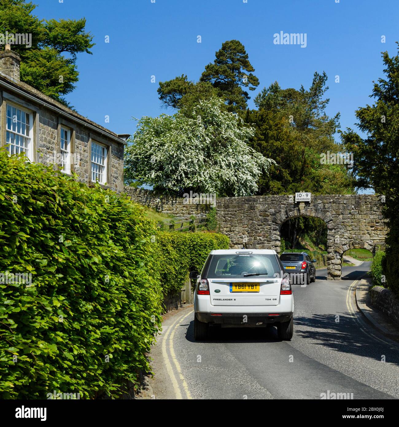 2 cars on country lane approaching narrow stone archway (arches, headroom warning sign 10' 9'')  - B6160, Bolton Abbey village, Yorkshire, England, UK Stock Photo