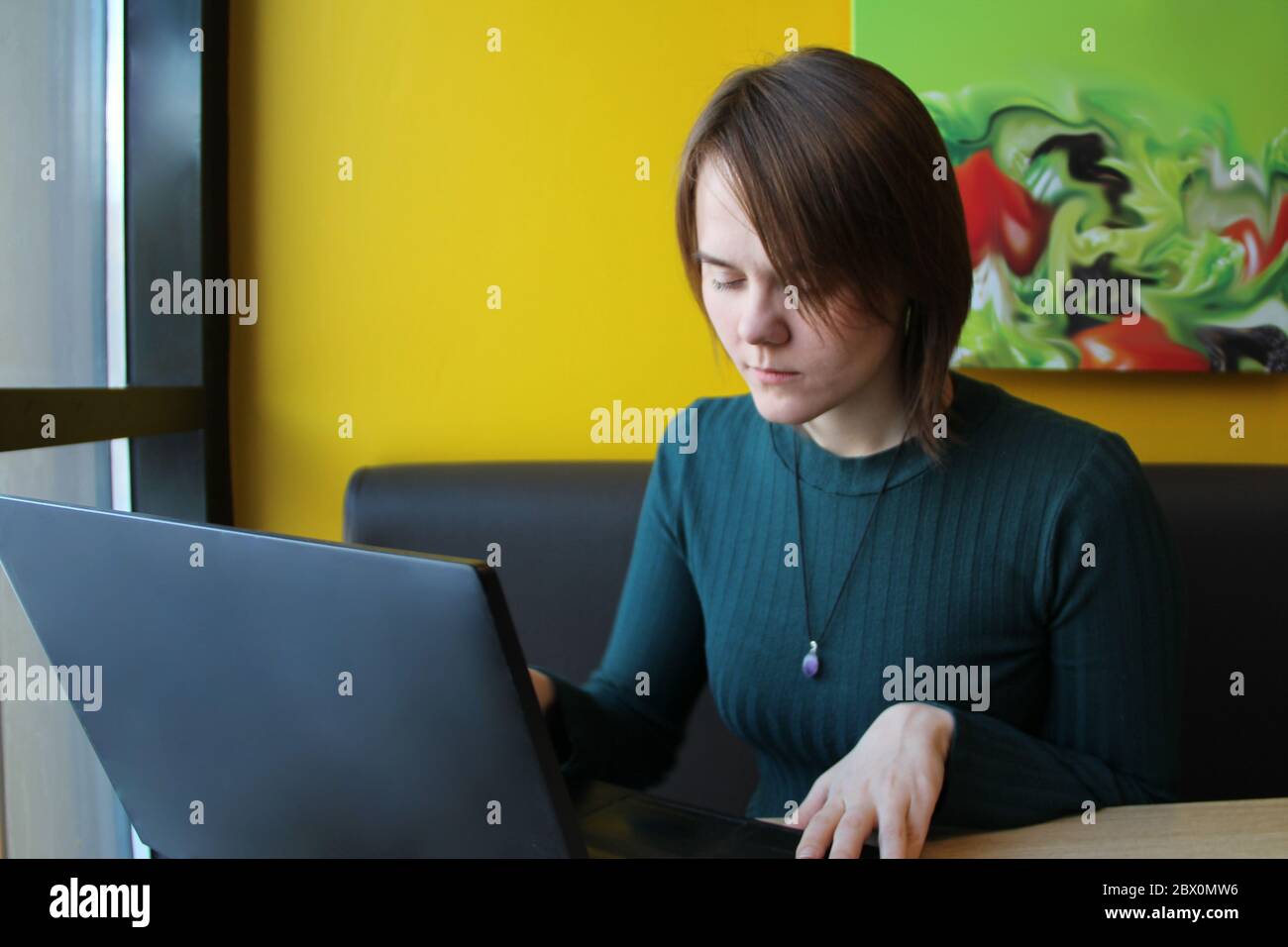 A girl with a calm expression sits, works at a laptop at a table in a cafe on a brown sofa against a yellow wall. He looks at the computer screen with his eyes closed. Stock Photo
