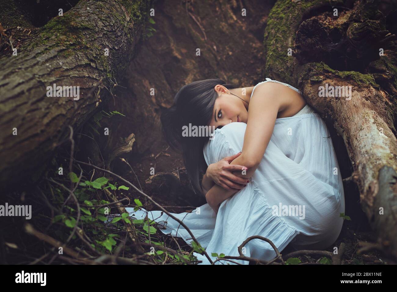 Beautiful woman among dark tree roots looks into camera . Mother earth embrace Stock Photo