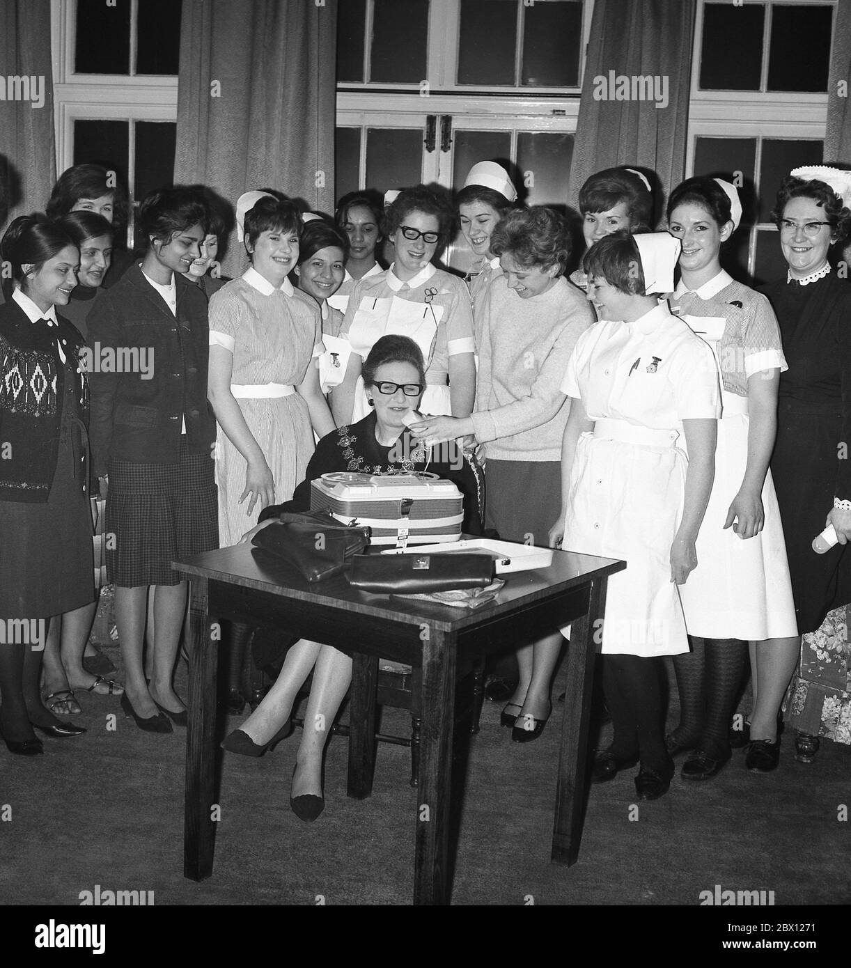 1960s, historical, at a hospital nursing reception, a group of female nurses standing behind the local Lady Mayoress of Lewisham who is sitting at a table in the room, being interviewed by a lady using an audio reel to reel tape player, South London, England, UK. Stock Photo