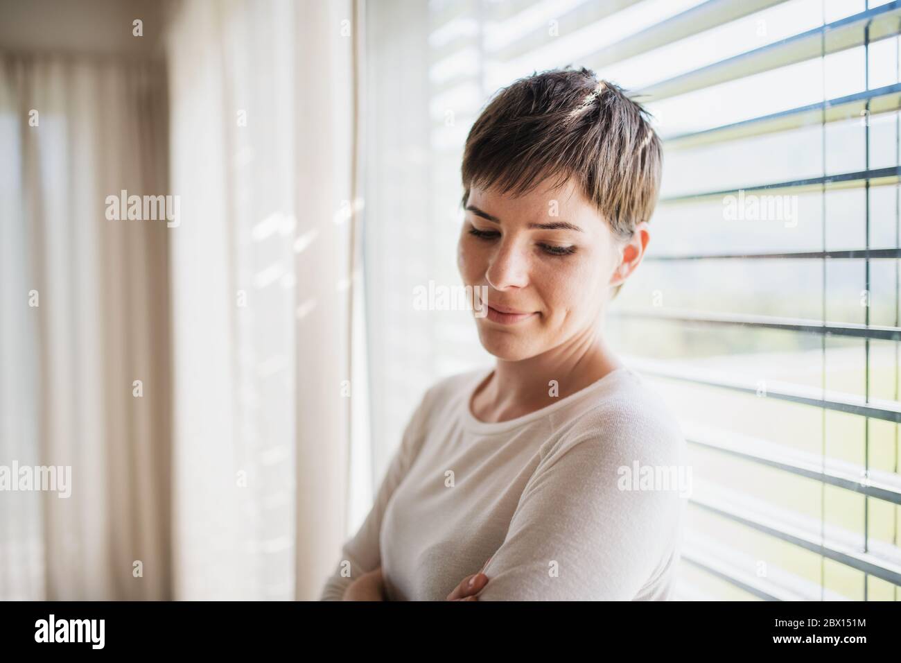 Portrait of young woman standing by window indoors at home, daydreaming. Stock Photo