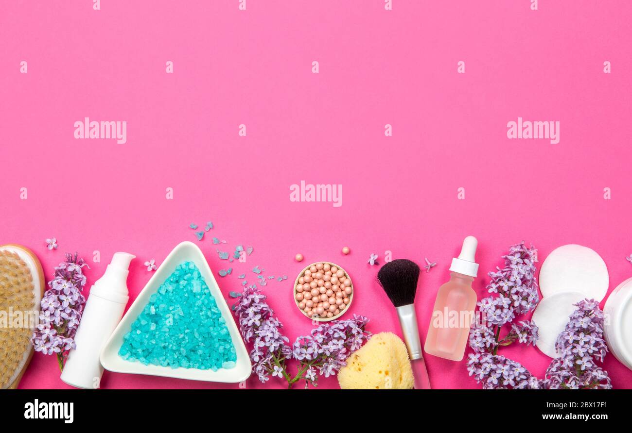 Flat lay view of various white cosmetic and beauty products containers with lilac (Syringa) blossoms on pink studio background. Lower border. Stock Photo