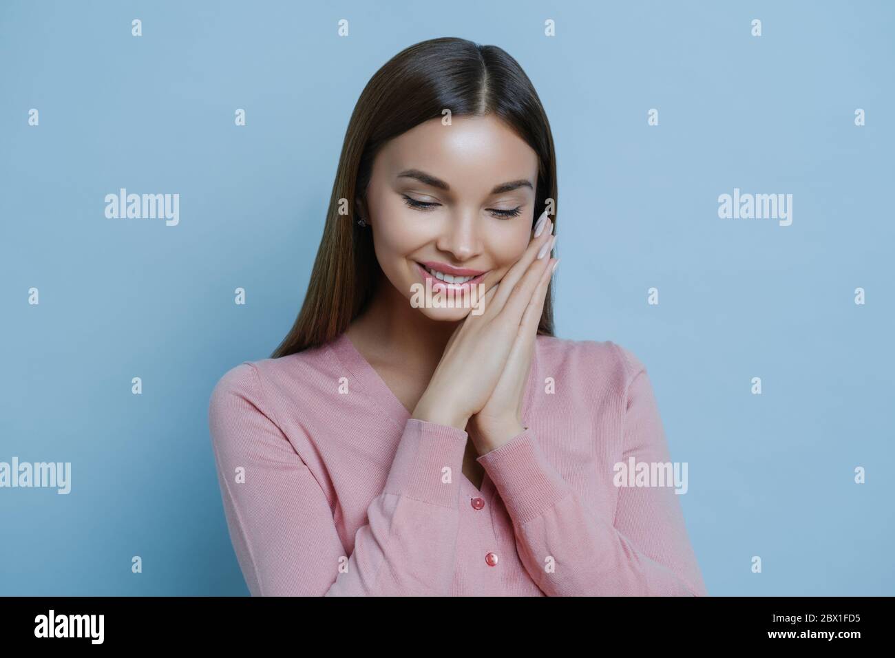 Portrait of glad shy woman keeps hands pressed together near face, concentrated down, pleased to hear compliment, has charming expression, has flirtin Stock Photo