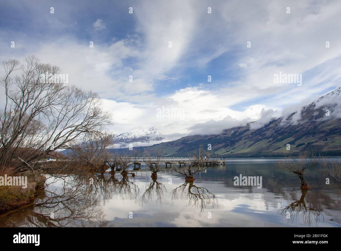 The Willows of Lake Wakatipu and Glenorchy Wharf, Otago, New Zealand. Magical scene in winter with cloudy sky, mountain and trees reflected in water Stock Photo