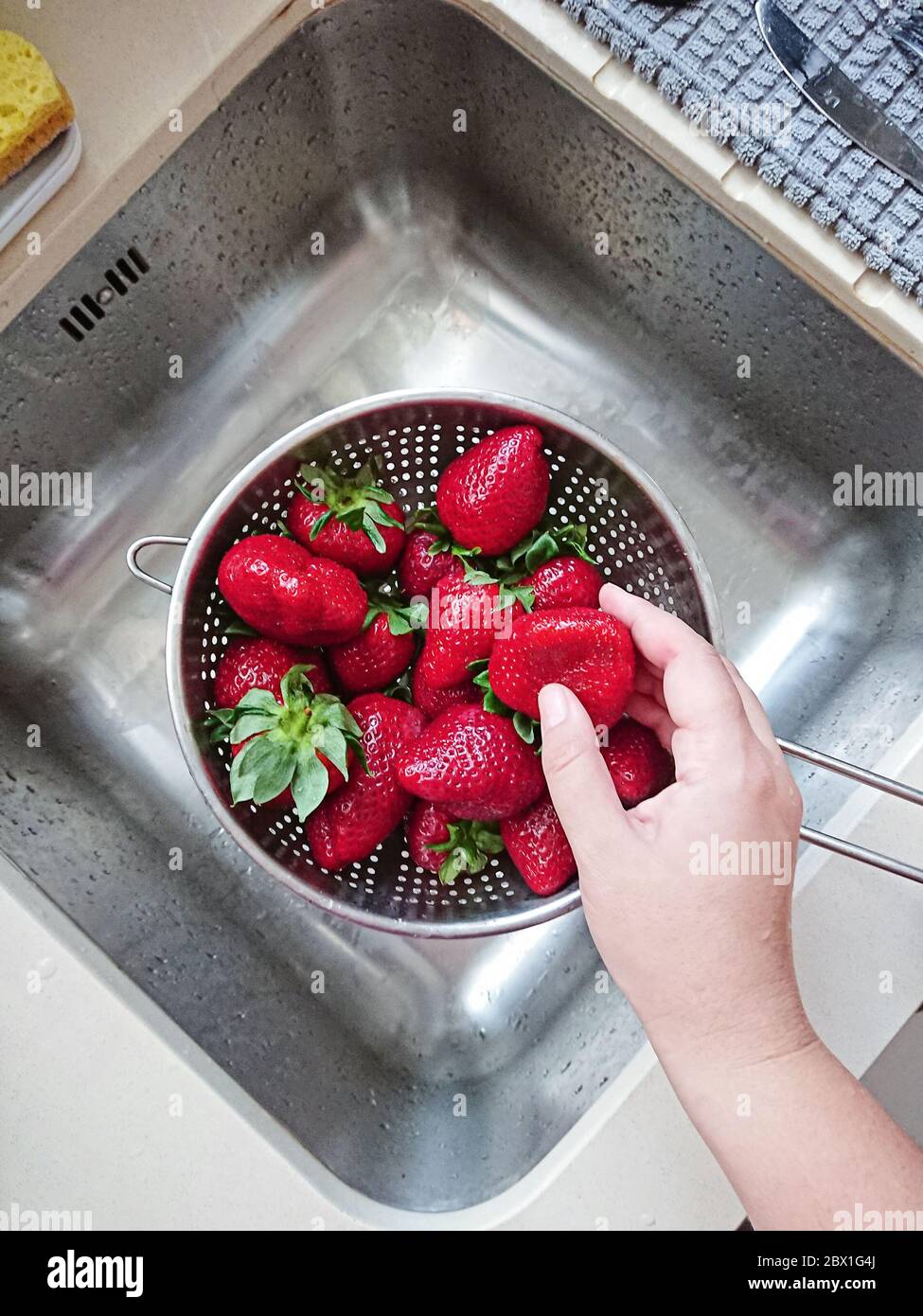 Washed ripe juicy red strawberries in mesh strainer above sink in kitchen, POV and top view Stock Photo