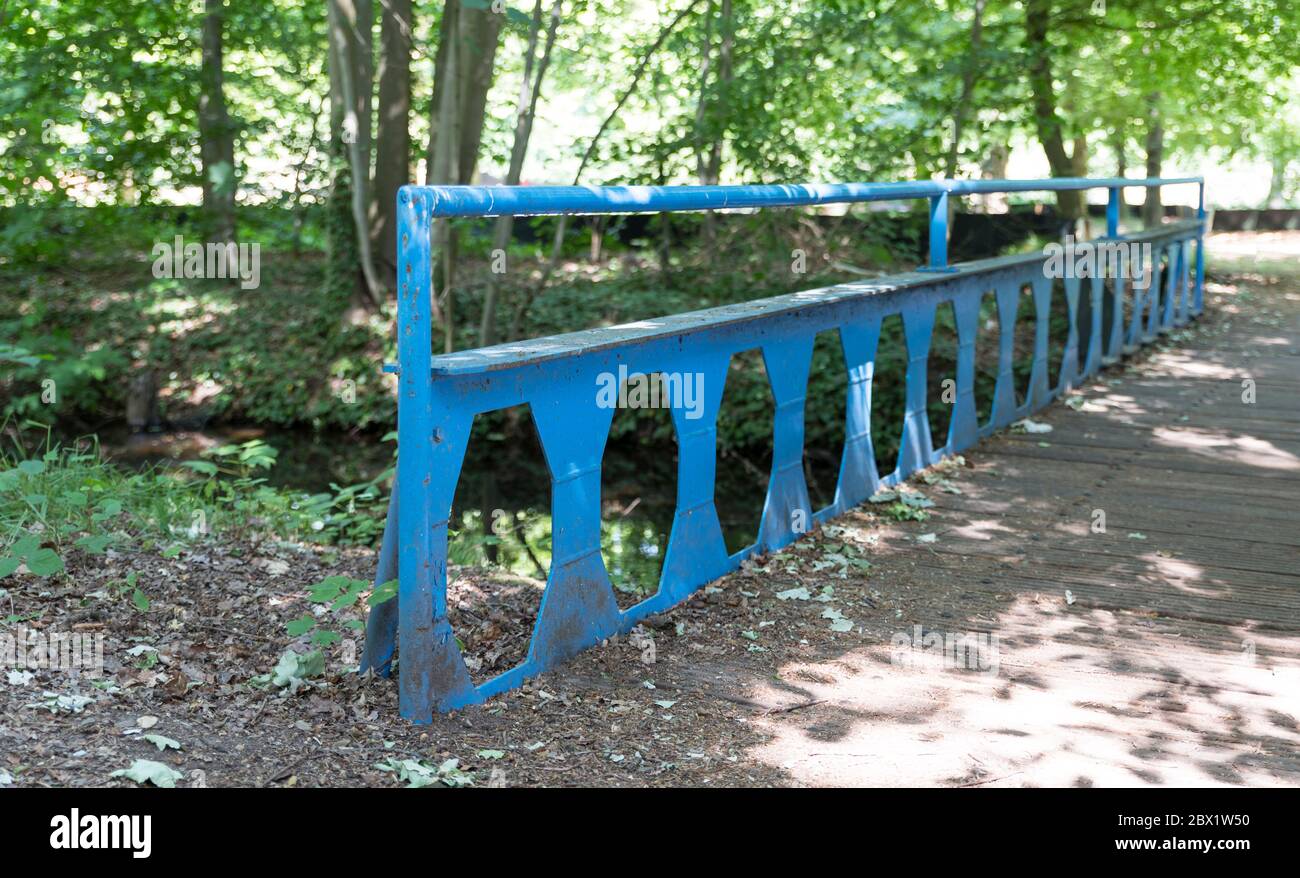 old blue metal bridge with wooden planks over a small river in a park with lots of greenery, trees and walking path Stock Photo