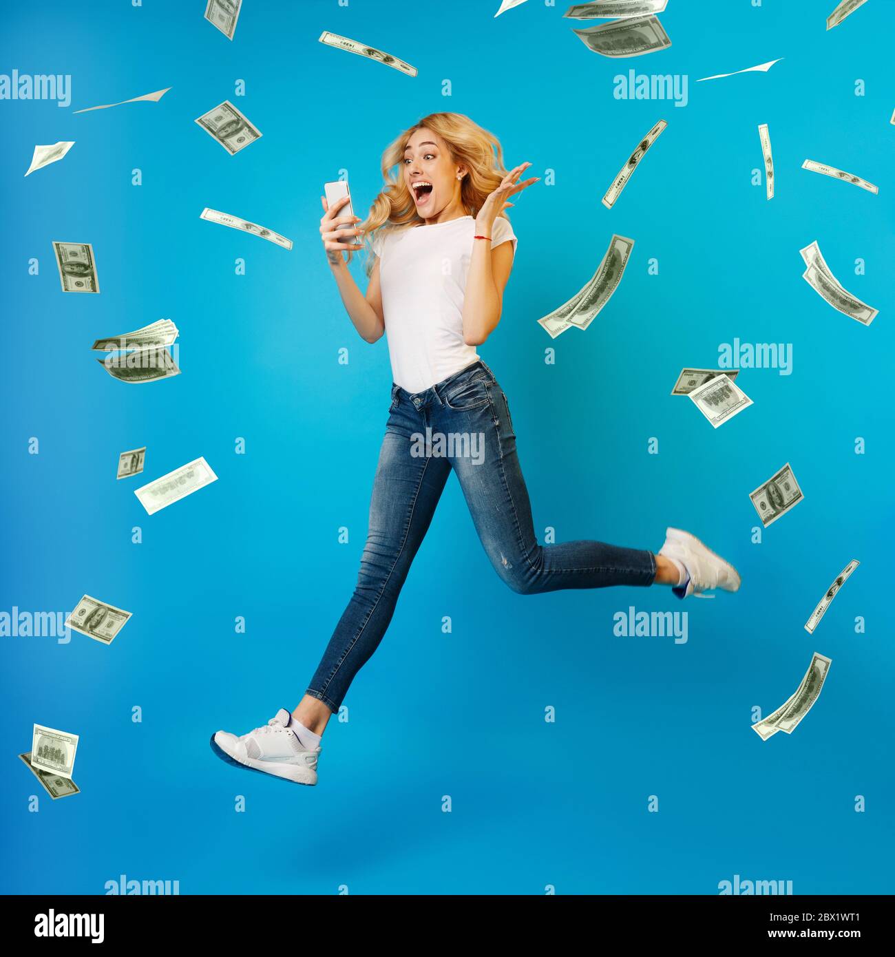 Online Lottery. Overjoyed Woman Jumping With Smartphone Under Money Shower, Celebrating Victory Stock Photo