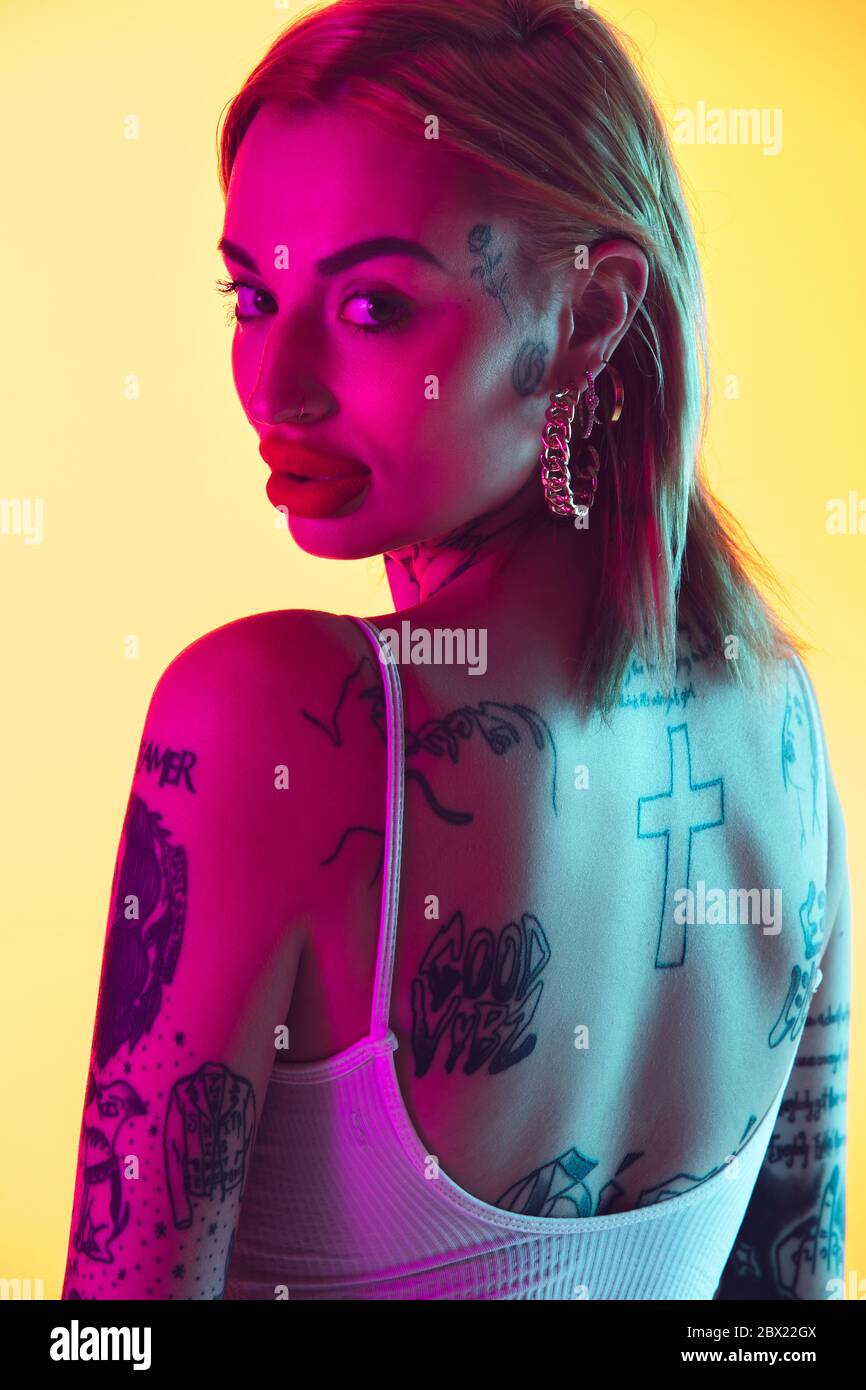 Close up young caucasian woman in swimsuit posing on gradient yellow studio background in neon light. Beautiful model with tattoos. Human emotions, sales, ad concept. Resort and vacation, summertime. Stock Photo