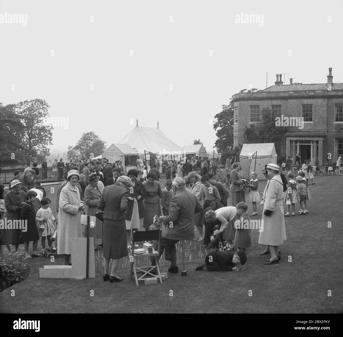 1950s, historical, adults and children on the lawns at a traditional English Country House garden party or fete, with some mothers snd children queuing up to play a game, England, UK. Perhaps the heyday of the 'fete', was in this era when a large country house of the village beionging  to a local landowner or squire would hold in the grounds a garden party for local people to come together raise monies for good causes in their community. Stock Photo