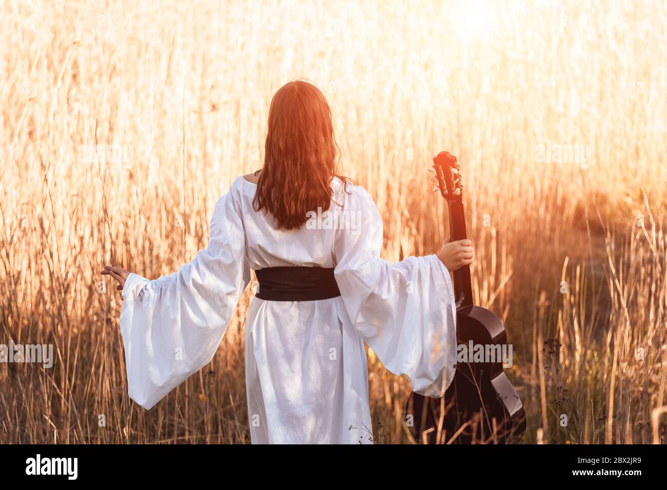Back view of slender young woman with red hair in a white medieval dress and belt holding acoustic guitar and walking through the sunset light field. Stock Photo