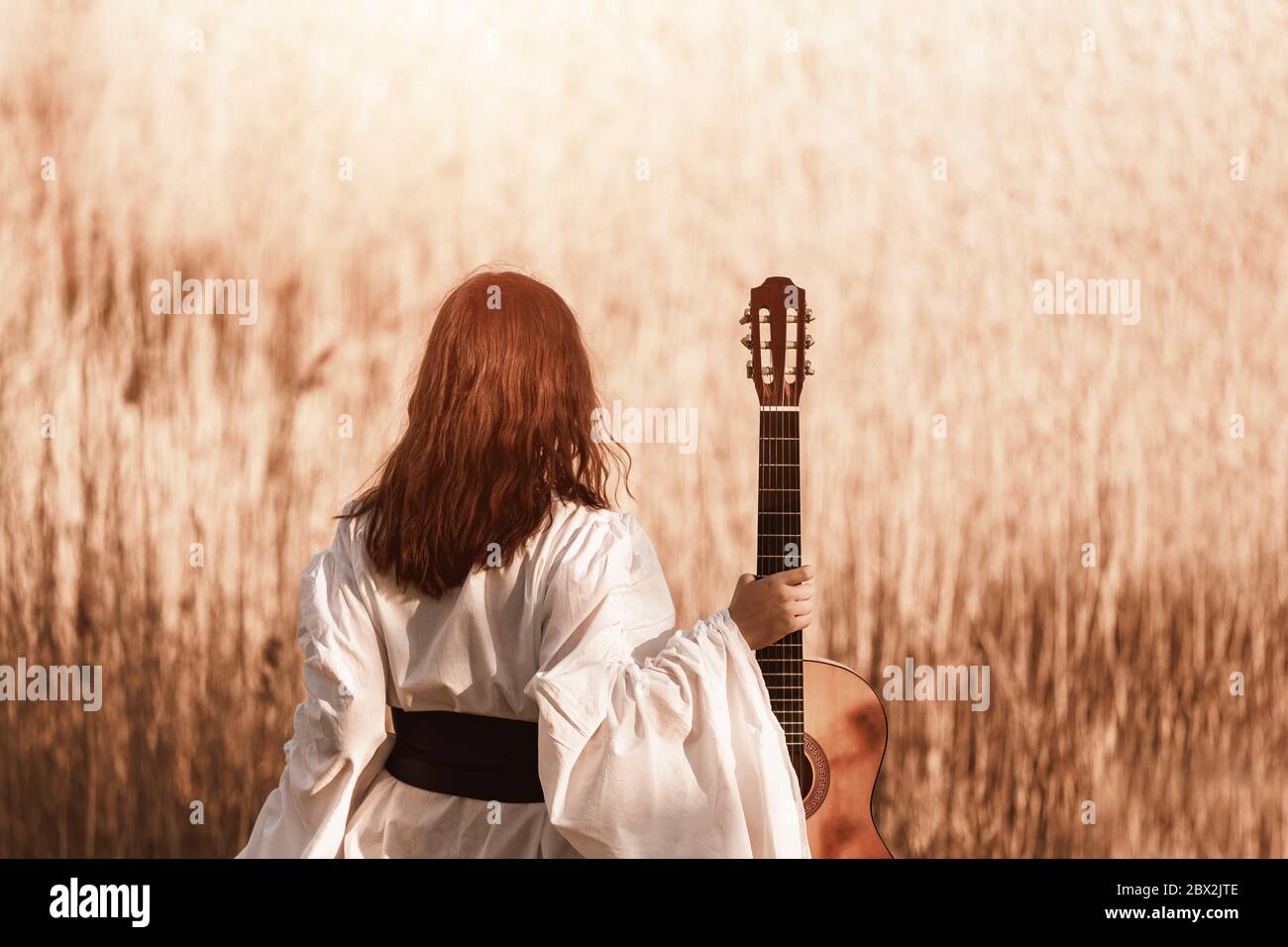 Beautiful young woman with red hair in a white medieval dress holding guitar and walking through the sunny field at warm light of sunset. Lens flare Stock Photo